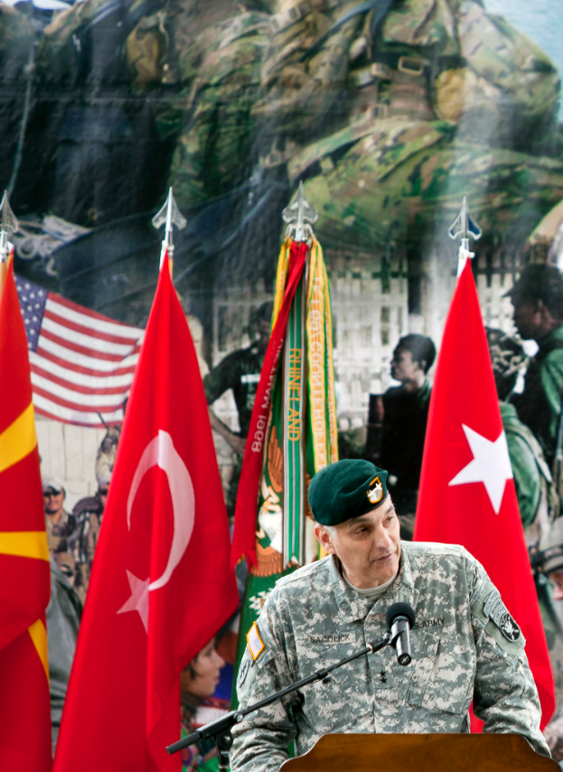 Ein Soldat steht an einem Rednerpult mit einem Mikrofon, umgeben von verschiedenen Flaggen und einem Plakat mit Militärpersonal im Hintergrund.