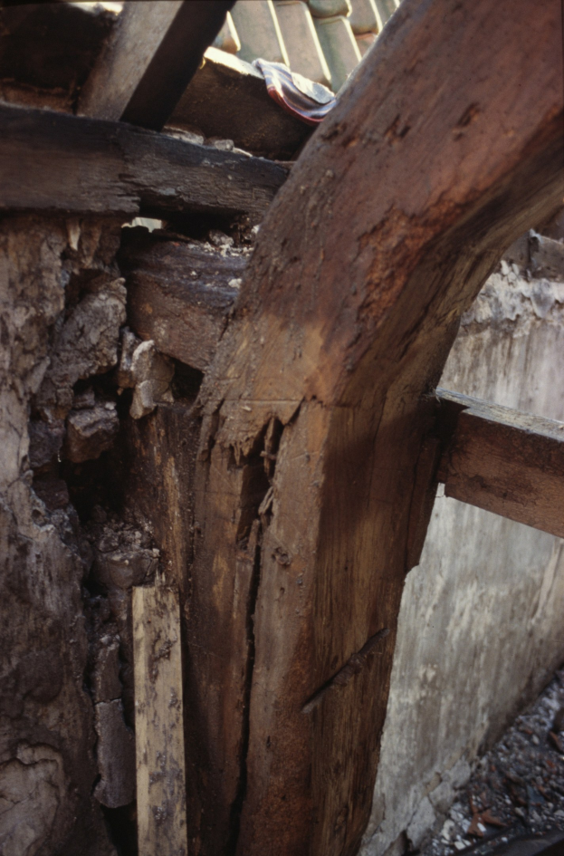 Close-up of a wooden beam on a building's side, with a wall in the background and a roof above, showing signs of repair.
