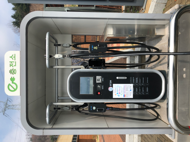 Eine Ladestation für Elektrofahrzeuge mit einer Maschine, Kabeln und einer Texttafel, vor dem Hintergrund von Gebäuden, Bäumen, Polen, Kabeln und dem Himmel.