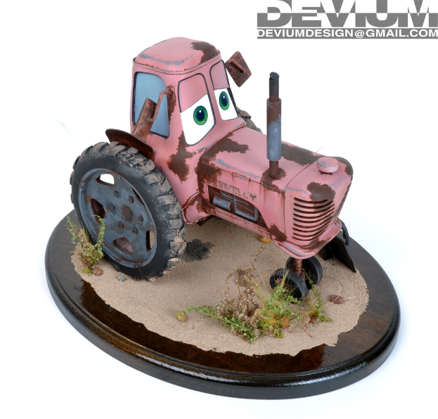 Ein Spielzeugtraktor auf einer braunen, sandigen Oberfläche mit spärlicher Vegetation und einem Wasserzeichen im oberen rechten Eck.