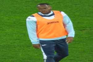 Ein Fußballspieler in einem orangen T-Shirt und blauen Hosen, der entschlossen über ein grünes Feld läuft.