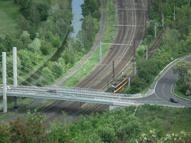 Ein Zug fährt über eine Brücke über einen Fluss, mit Fahrzeugen auf der Straße darunter und Strommasten mit Drähten, die entlang der Brücke verlaufen, vor einem Hintergrund aus Bäumen und Gras.