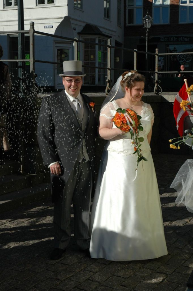 Ein Paar steht vor einem Zaun und einem Gebäude, wobei die Frau einen Strauß hält und verschiedene Objekte in der rechten Ecke zu sehen sind.