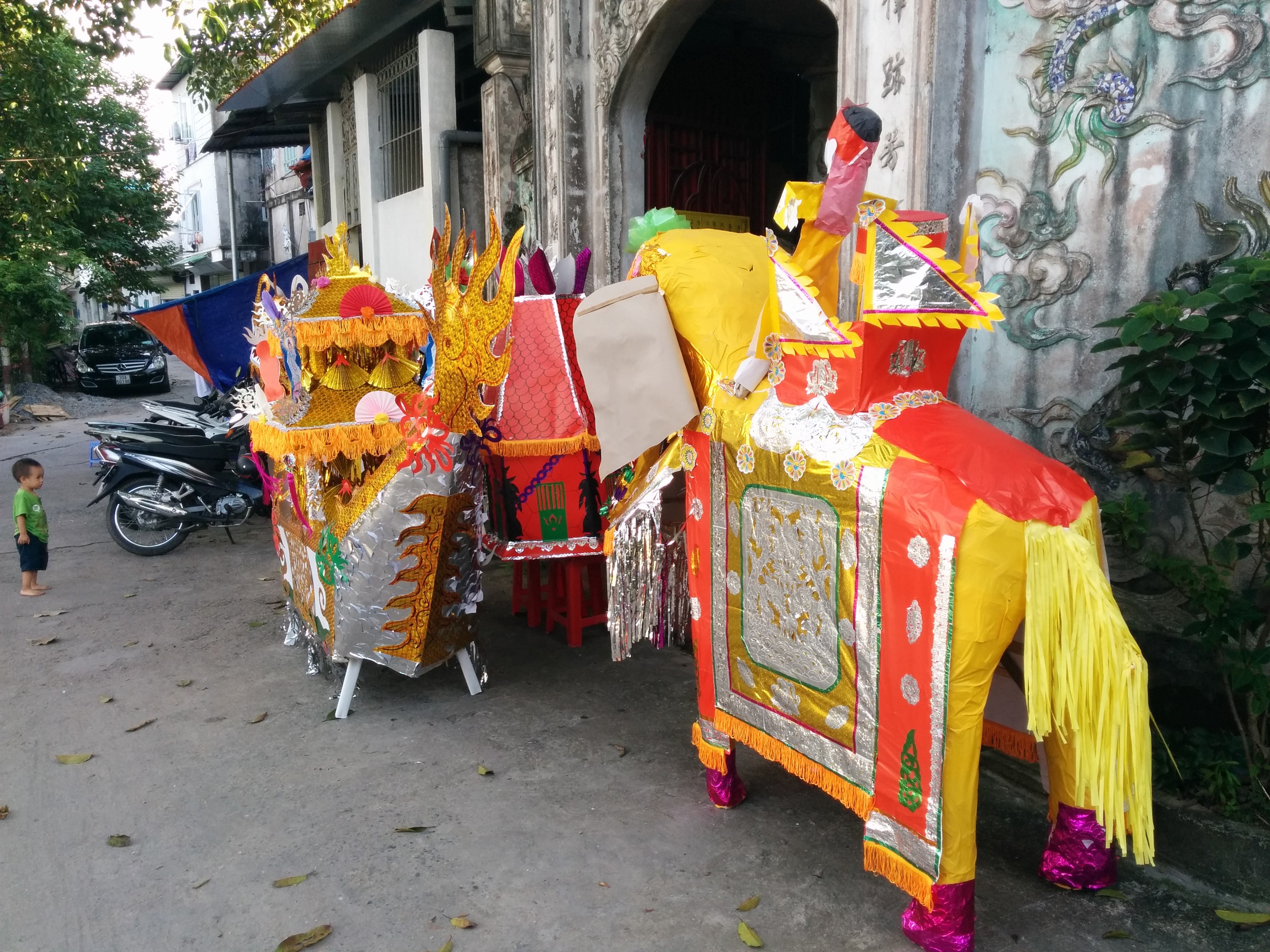 A vibrant Chinese New Year parade scene in Ho Chi Minh City with vehicles, a person on the left, buildings and trees in the background, a painted wall on the right, and decorative parade items in the foreground.