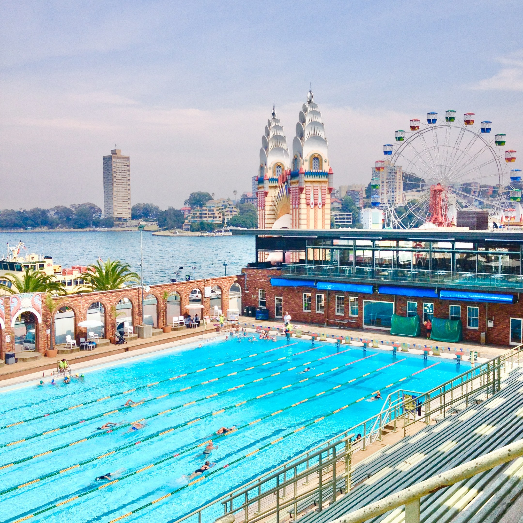 Large swimming pool with people swimming, surrounded by railings and benches, with a ferris wheel, buildings, trees, and a cloudy sky in the background.
