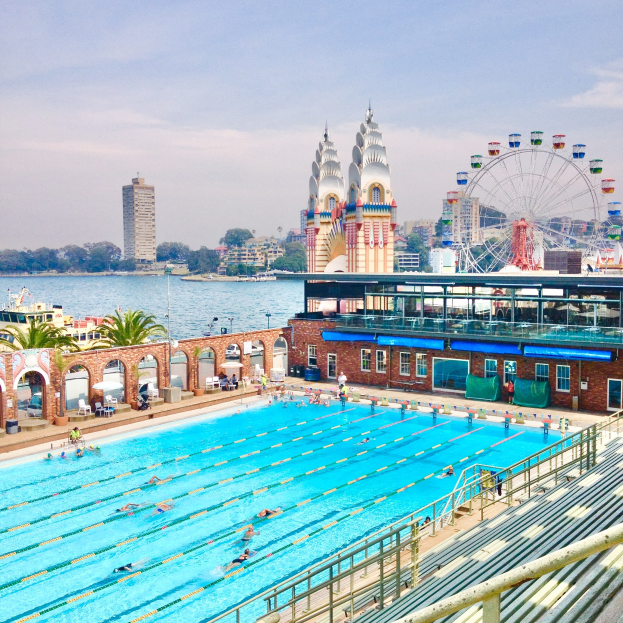 Large swimming pool with people swimming, surrounded by railings and benches, with a ferris wheel, buildings, trees, and a cloudy sky in the background.