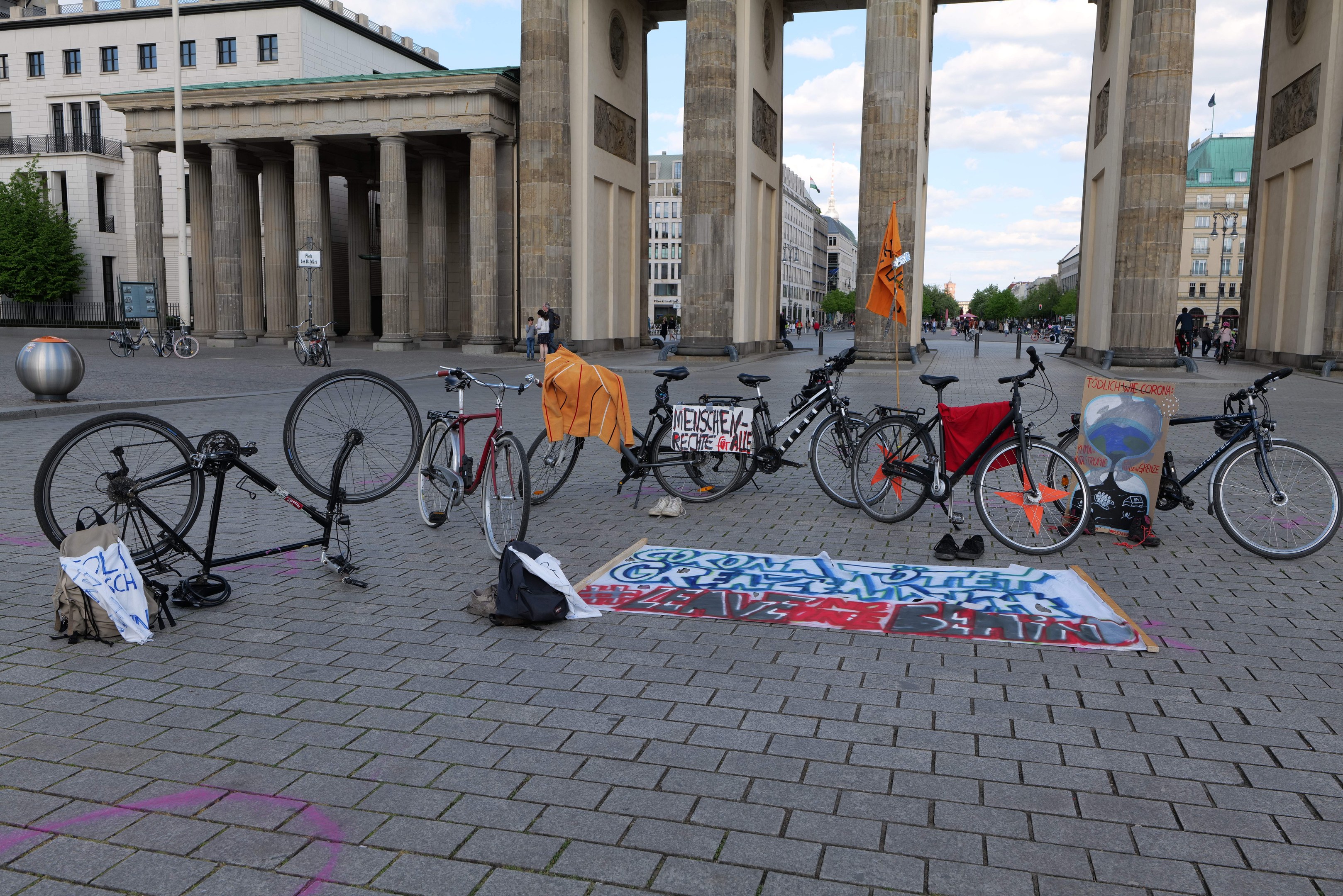 Bicycles parked in front of the Brandenburg Gate in Berlin, Germany, with bags and a banner nearby, set against buildings, trees, and a clear blue sky.