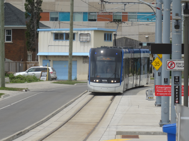 Eine Straßenbahn fährt durch eine Straße mit hohen Gebäuden, mit Masten, Schildern, einem Müllcontainer, einem Auto und einem Baum.