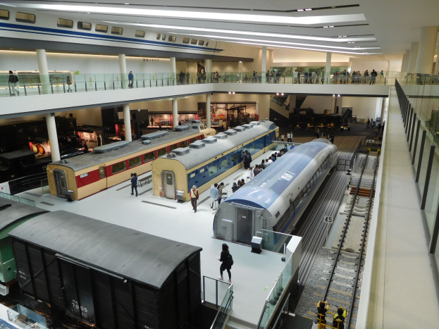 Eine Museumsausstellung, die verschiedene Züge in unterschiedlichen Größen und Farben zeigt, mit Menschen, die vom Boden, Geländern und Säulen aus beobachten.