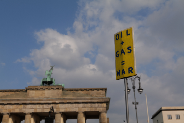 Gelbes Schild mit der Aufschrift "Öl und Gas Krieg" im Vordergrund mit dem Brandenburger Tor in Berlin, Deutschland, und Gebäuden, Masten, Lichtern und einer Statue im Hintergrund unter einem bewölktem Himmel.