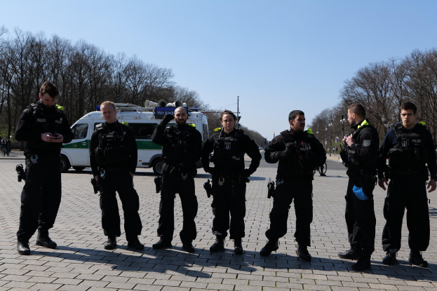 Eine Gruppe von Polizisten steht vor einem Rettungswagen auf einer von Bäumen gesäumten Straße unter einem klaren Himmel.