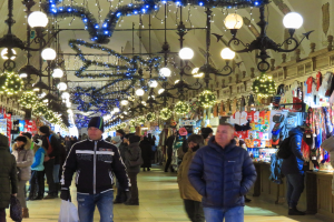 A bustling Christmas market with people walking, some wearing caps and holding bags, under hanging lights and decorations, with clothing and goods displayed on the right and a wall in the background.