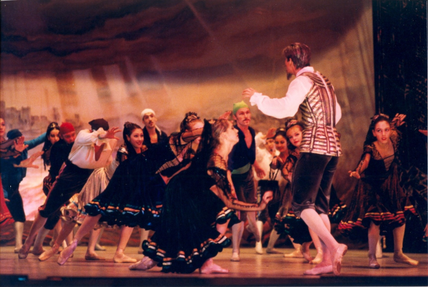 Eine Gruppe von Menschen in bunten Kostümen tanzt auf einer hell erleuchteten Bühne mit einer Wand im Hintergrund.