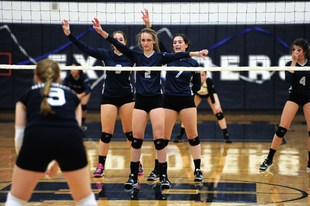 Drei Mädels in einem Volleyballspiel, die auf dem Feld stehen und die Hände heben, mit einem Netz davor und einem Zaun im Hintergrund.