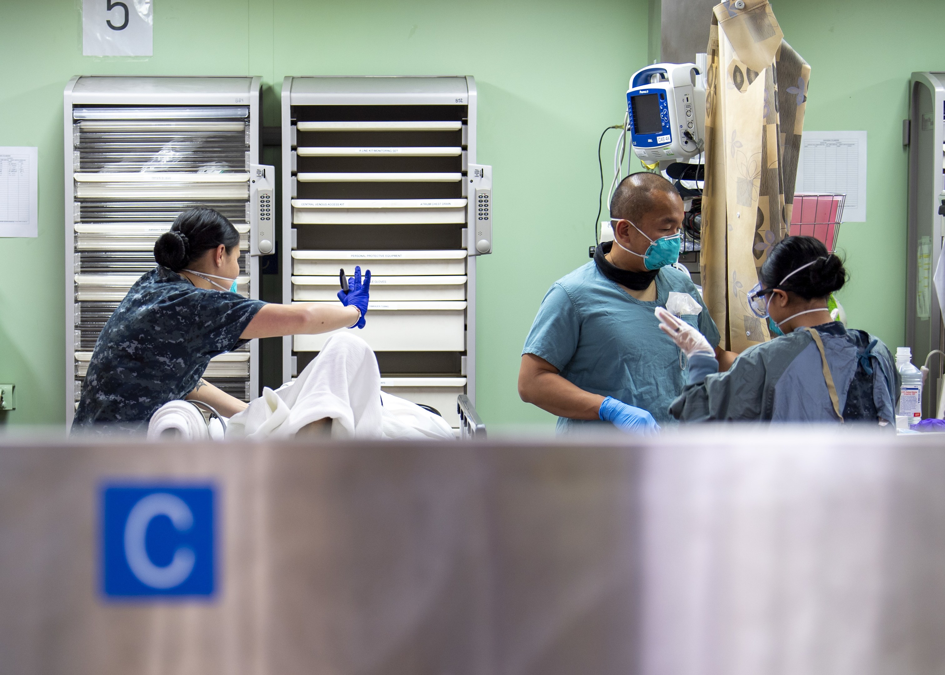 Medical professionals in scrubs and masks attending to a patient on a bed in a hospital room with medical equipment and supplies in the background.