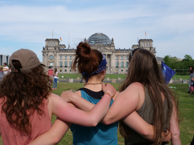 Drei Frauen in verschiedenen farbigen Kleidern, eine trägt eine Mütze, stehen vor dem Reichstagsgebäude in Berlin, Deutschland, mit Menschen, Fahnen, Bäumen und Wolken im Hintergrund.