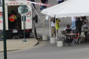 Ein Krankenwagen befindet sich auf der linken Seite einer Straße, während auf der rechten Seite ein Zelt viele Menschen schützt, mit einem Tisch und einem Stuhl in der Nähe.