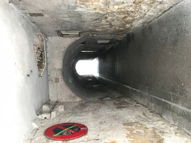 Ein Tunnel mit einem roten und schwarzen "Eintritt verboten"-Schild auf dem Boden, beleuchtet von Lampen öber ihm, das darauf hinweist, dass es Teil eines Abwassersystems ist.