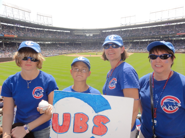 Drei Frauen mit Mützen und Brillen, ein Junge mit Brille und Mütze, der einen Ball und ein Schild hält, stehen in einem Stadion mit Publikum und Feld dahinter, unter einem Himmel mit einigen Lichtern.
