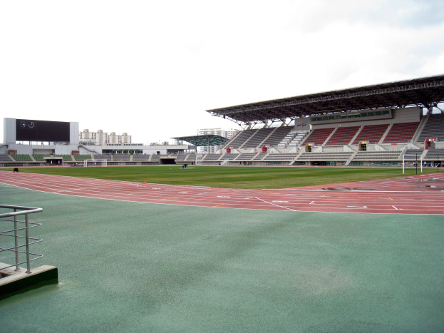 Großes Stadion mit zentraler Laufbahn, linker Begrenzung, fernen Gebäuden und einem klaren blauen Himmel.