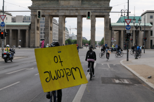 Eine Gruppe von Radfahrern mit Helmen fährt vor dem Reichstagsgebäude in Berlin, Deutschland, vorbei, wobei einer ein gelbes Schild h"alt, Lichtmasten, Verkehrszeichen, Geb"ude, B"ume und ein klarer blauer Himmel im Hintergrund zu sehen sind.
