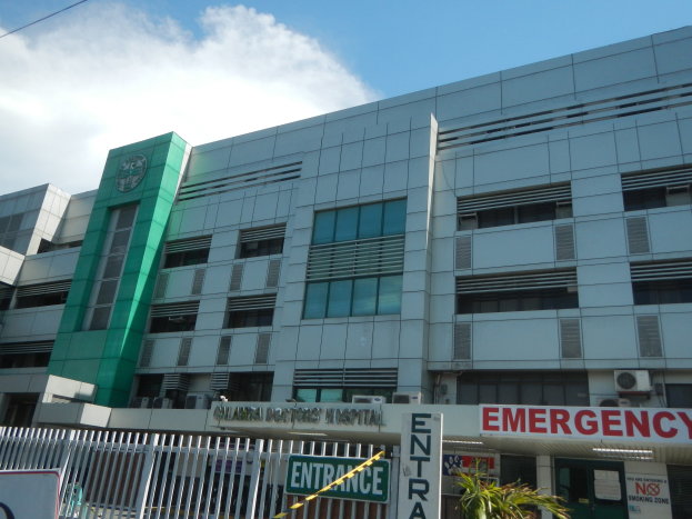 Großes Gebäude mit einer "Notfall"-Schilder, umgeben von einem Metallzaun, umgeben von Pflanzen, Informationsschildern, Fahrzeugen und Kabeln unter einem bewölkten Himmel.