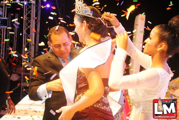 Ein Mädchen auf der Bühne bei einer Miss World Show mit einer Krone, dahinter ein Mädchen, das die Krone anpasst, und ein Mann in einem schwarzen Anzug, der vor ihr die Hand schüttelt, vor einem dunklen Hintergrund mit Metallrahmen.