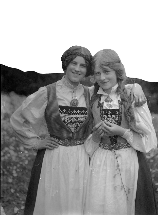 Zwei junge Frauen in traditioneller bayrischer Tracht, mit weißen und schwarzen Kleidern mit aufwendiger Stickerei und Perlen, posieren vor einer Kulisse aus üppigen grünen Pflanzen und einem hellen weißen Himmel.