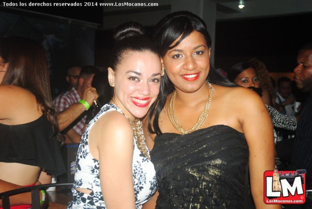 Zwei Frauen in modischer Kleidung stehen und lächeln in einer Bar, während andere Menschen hinter ihnen tanzen und eine Lampe an der Decke leuchtet.