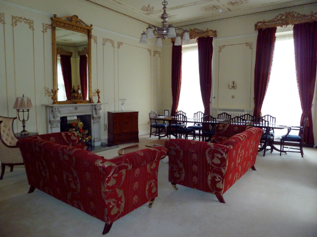 Zwei rote Sofas in einem Wohnzimmer, ein kleiner Teetisch mit einem Buch darauf, ein wandmontierter Spiegel, eine Lampe auf einem Tisch, ein Blumenbouquet vor einem Kamin, ein Esstisch mit Stühlen, von der Decke hängende Lampen, Vorhänge an einer Stange und ein leerer Stuhl.