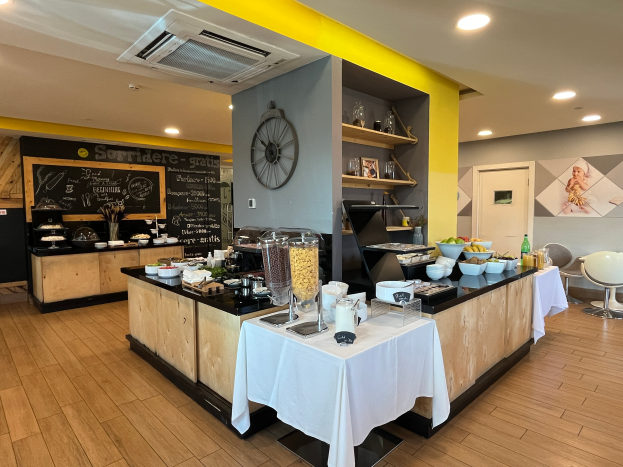 Ein Restaurant-Innenraum mit Tischen und Stühlen, Speisen, einer Tafel mit Text, einer Wanduhr, Deckenleuchten, einer Klimaanlage und einer Tür im Hintergrund, das sich im Hôtel de Ville in Paris befindet.