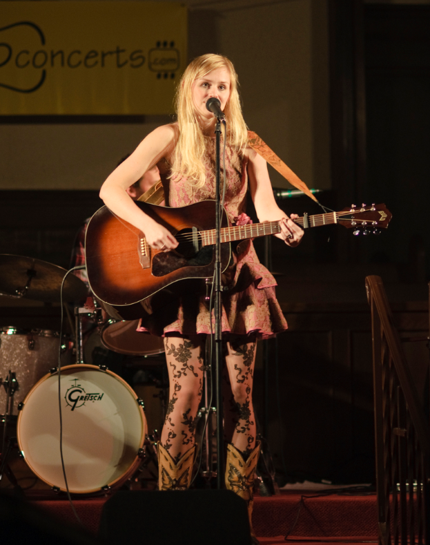 Eine Frau spielt Gitarre und singt auf der Bühne bei einem Konzert, mit einer Band und einem Bildschirm im Hintergrund.