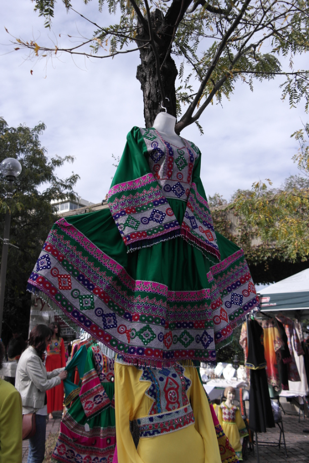 Eine Schaufensterpuppe steht in der Mitte eines belebten Einkaufsgebiets mit mehreren Geschäften, einer Person, die Kleider an einem Stab arrangiert, einem Gebäude und Bäumen im Hintergrund.
