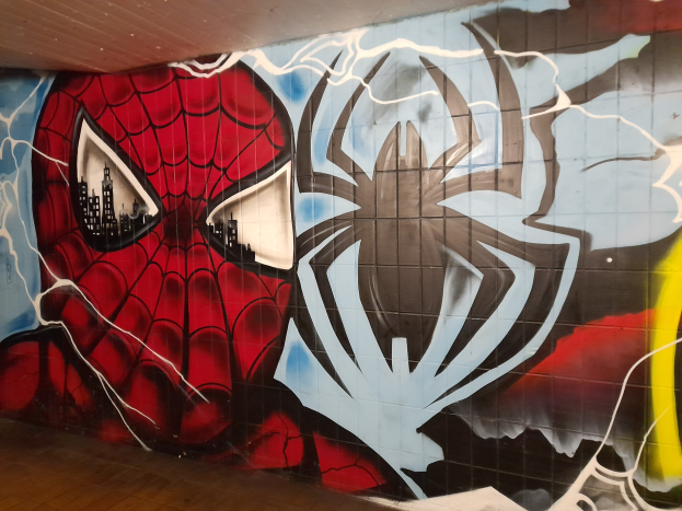 Buntes Wandgemälde von Spider-Man in rot und blauem Kostüm mit Webmuster und großem Spinnenlogo auf der Seite eines Gebäudes, das ihn mit weit aufgerissenen Augen und leicht geöffnetem Mund zeigt.