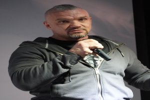 Ein Mann in einer schwarzen Kapuzenjacke und Anzug, der ein weißes Hemd und zurückgekämmtes Haar trägt, hält ein Mikrofon mit einem selbstbewussten Gesichtsausdruck vor einer Wand.