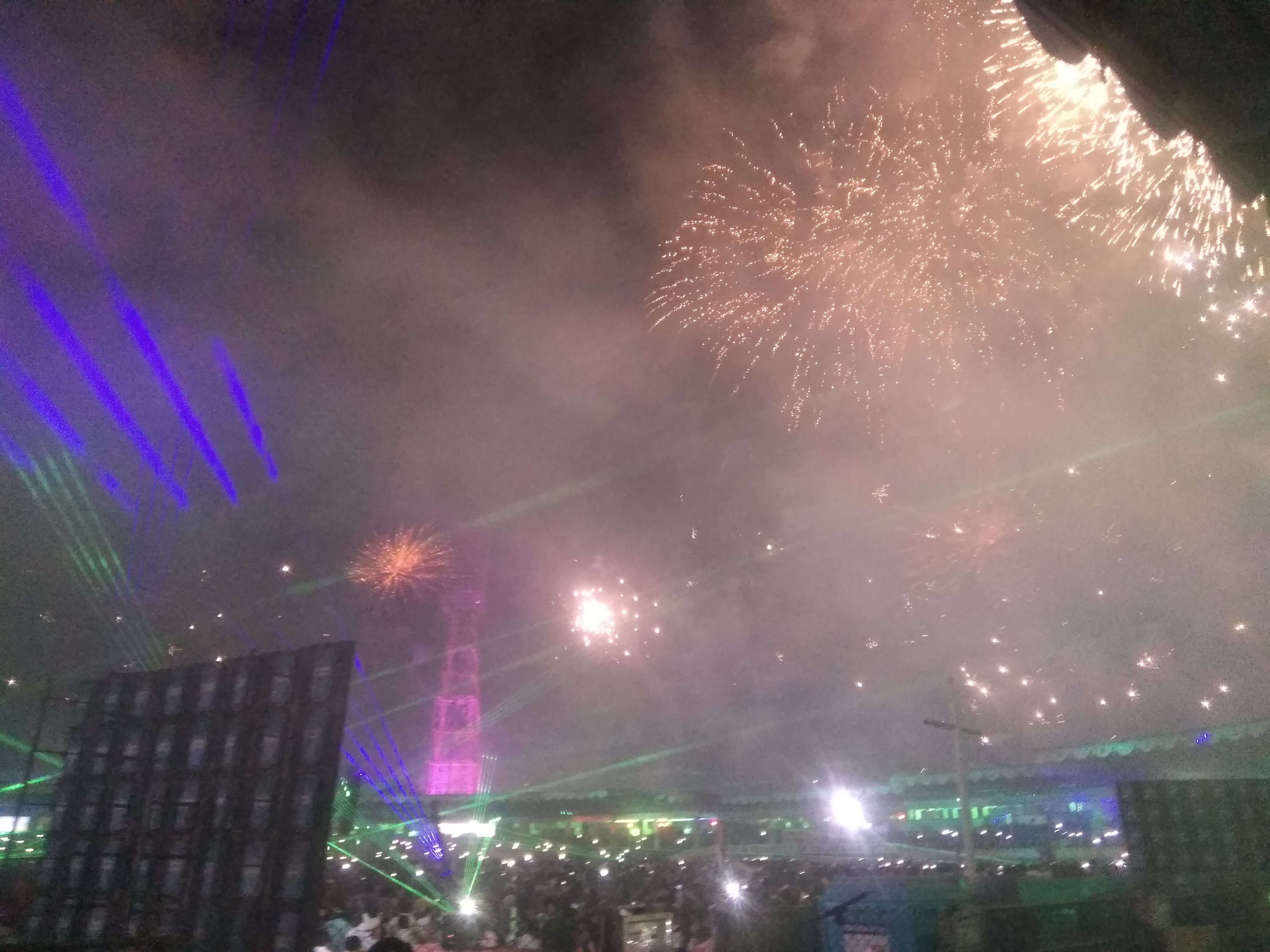 Eine Menschenmenge bei einem Konzert mit beleuchteten Gebäuden und Pfählen im Hintergrund, unter einem Himmel voller farbiger Feuerwerke.
