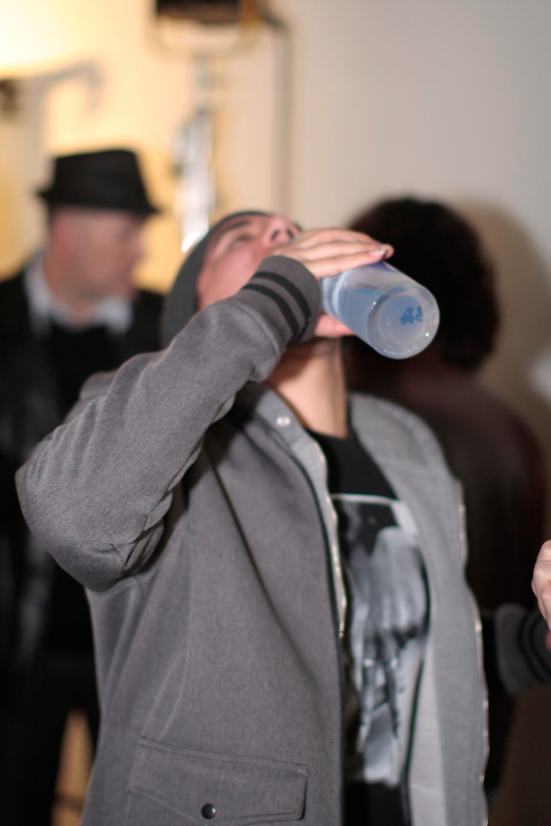 Ein Mann in der Mitte des Bildes hält eine Flasche und trinkt Wasser, mit ein paar anderen Menschen im Hintergrund sichtbar.