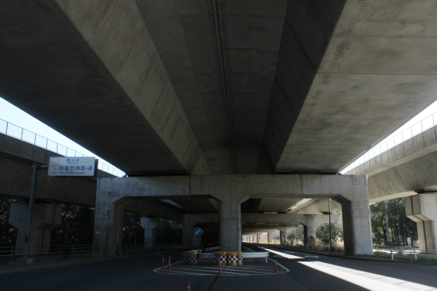 Autobahn unter einer Brücke mit Absperrkegeln, Barrieren und Texttafeln am Straßenrand, flankiert von Geländern, Bäumen und einem klaren blauen Himmel im Hintergrund.