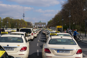 Eine lange Reihe von Taxis, die an der Seite einer belebten Straße in Berlin, Deutschland, geparkt sind, mit Fahrzeugen, Fahrradfahrern und Füülern, flankiert von Laternen und Bäumen, mit Gebäuden, einem Bogen und einem Turm im Hintergrund unter einem bewölktem Himmel.
