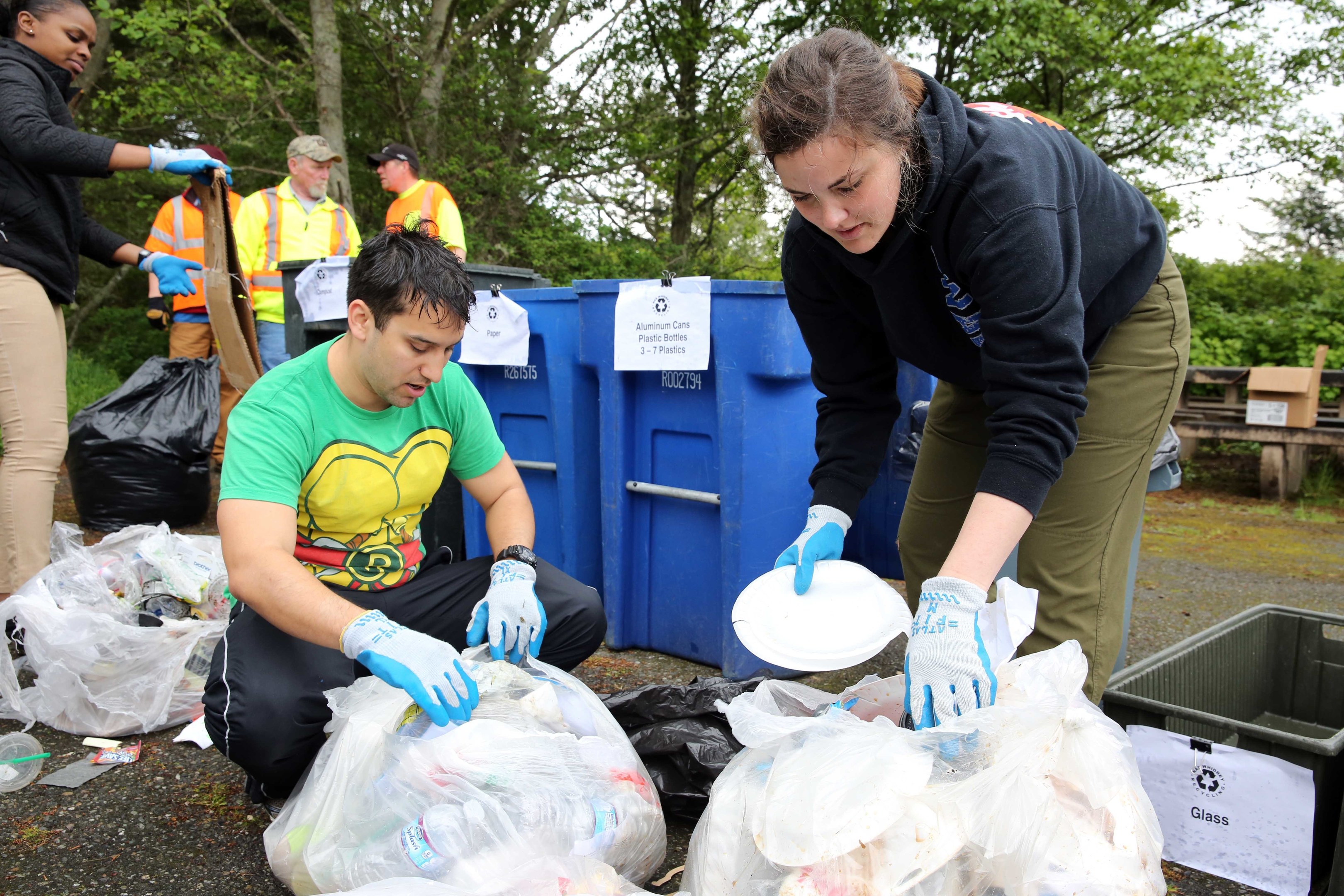 A group of people wearing gloves collect trash in a park, surrounded by discarded items, with a dustbin and bench nearby under trees and a clear sky.