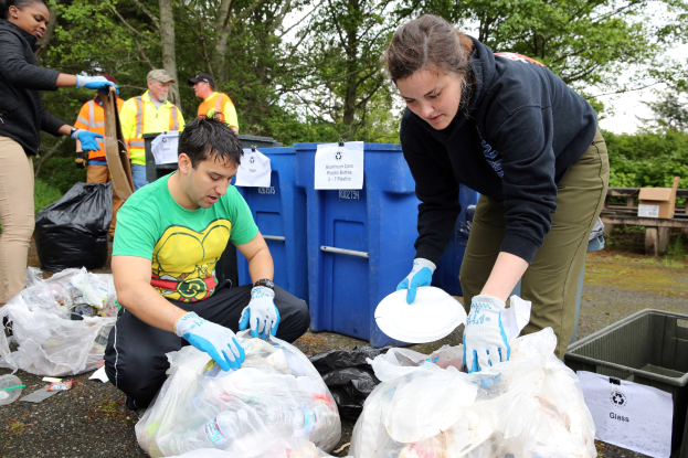A group of people wearing gloves collect trash in a park, surrounded by discarded items, with a dustbin and bench nearby under trees and a clear sky.