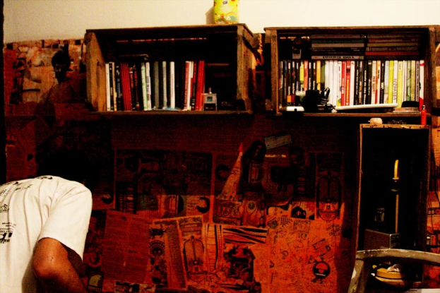 Eine Person steht vor einer Wand mit aufgeklebten Werbeplakaten, mit Schränken voller Fernbedienungen und Büchern im Hintergrund.