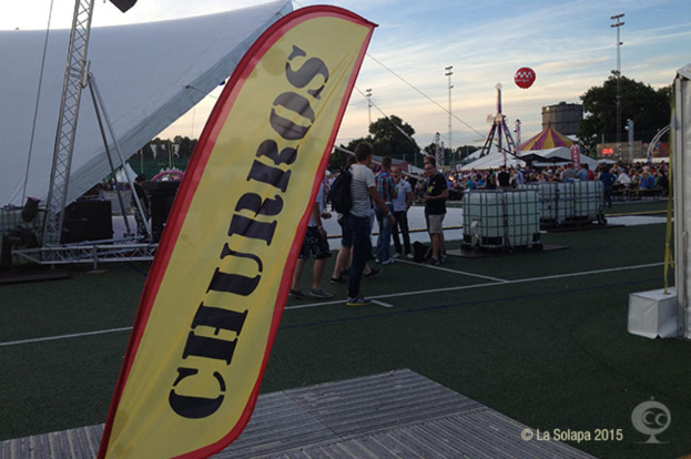 Eine KarnevalsSzene mit Zelten, einem zentralen Mast, Menschen, Bäumen, Strommasten, Straßenbahnen auf der Straße und einem Eisengestell auf der linken Seite unter einem HimmelHintergrund.