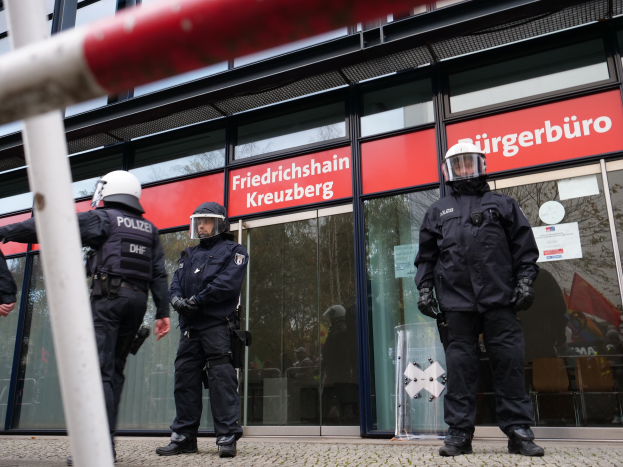 Eine Gruppe von Polizisten in Helmen und Handschuhen steht vor einem Gebäude mit Glaswänden und -türen, links ein Pfahl und im Hintergrund Schilder mit "Friedrichshain Kreuzberg" und Papiere an den Glaswänden geklebt.