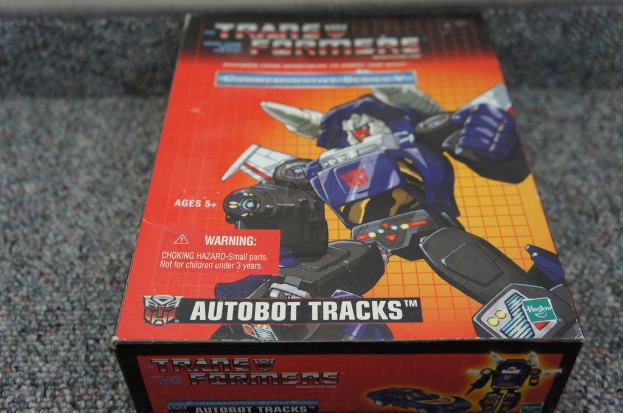 Ein roter, oranger und blauer Kasten mit einem Roboter und dem Text "AUTOBOT TRACKS" oben, vor einem unscharfen schwarzen Hintergrund.