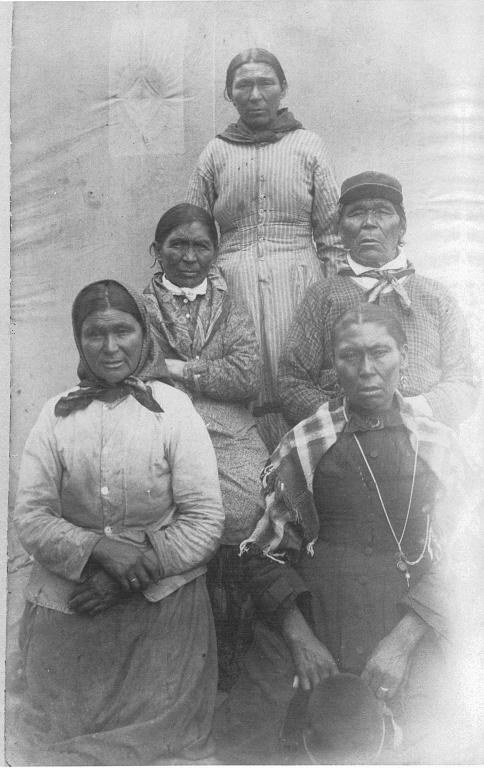 Schwarzes und weißes Foto einer Gruppe von indigenen Frauen, die in einer Reihe vor einer Wand stehen, einige tragen traditionelle Kleidung und Kopfschmuck, mit Text unten, der "Indigenous Women" lautet.