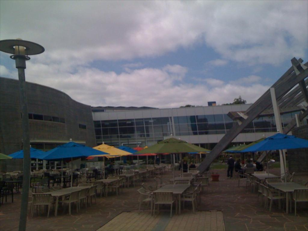 Eine Resort-Szene mit Stühlen, Tischen, Sonnenschirmen, einer Lampe im Vordergrund, Bäumen und Gebäuden in der Mitte, unter einem bewölkten Himmel.