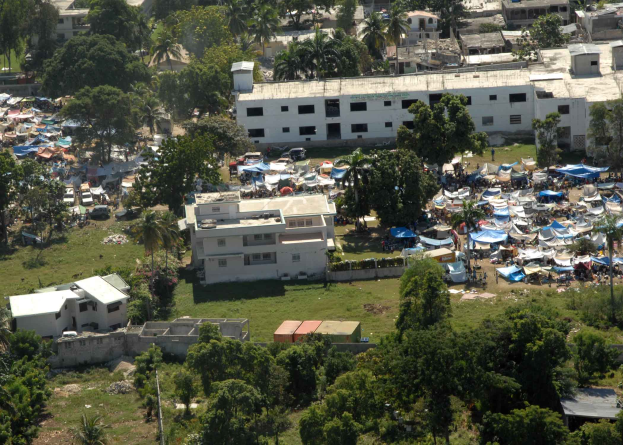 Eine Luftaufnahme einer großen Gruppe von Zelten in einer Stadt, umgeben von Gebäuden, Bäumen, Gras, Fahrzeugen und Polen, wahrscheinlich ein Flüchtlingslager.
