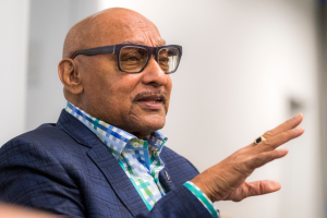 Ein Mann in Anzug und Brille spricht in ein Mikrofon und wendet sich an ein unscharfes Publikum.