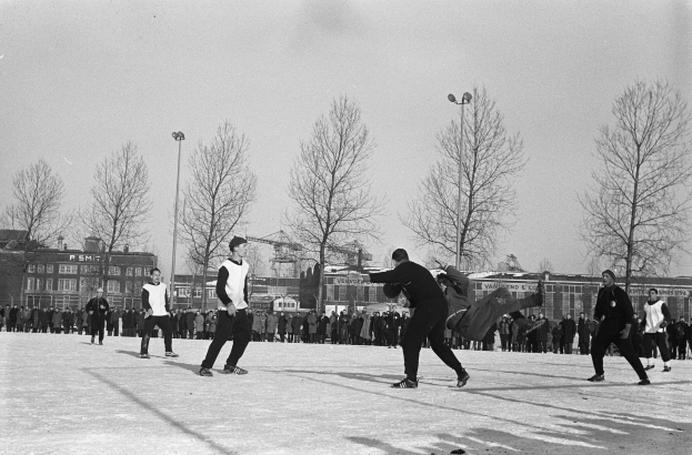 Schwarzes und weißes Bild von Menschen, die Eis Hockey im Schnee mit Bäumen, Gebäuden, Laternenmasten und einem klaren Himmel im Hintergrund spielen.